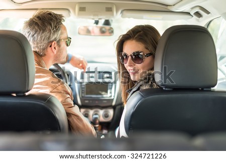 Rear view of a middle aged couple in a black seats car. They are about to leave for the weekend, wearing their leather coats and sunglasses. The smiling woman is looking at camera