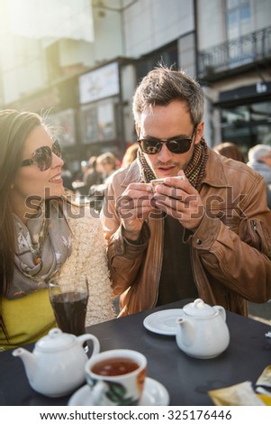 Backlit shoot. Trendy couple having fun and sitting at a terrace in the city center. The woman is wearing a woolen coat,The grey hair man has  a leather coat. A shopping bag is on the bar table