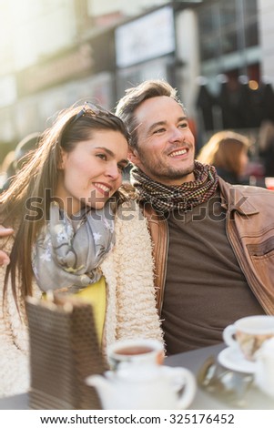 Trendy couple having fun and sitting at a terrace in the city center. The woman is wearing a woolen coat and a scarf The grey hair man has a beard and a leather coat A shopping bag is on the bar table
