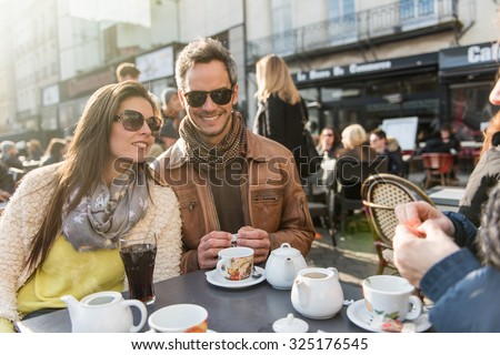 Backlit shoot. Trendy couple having fun and sitting at a terrace in the city center. The woman is wearing a woolen coat,The grey hair man has  a leather coat A shopping bag is on the bar table