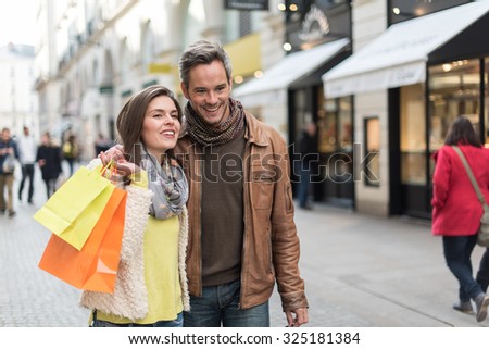 Trendy couple walking in a cobbled car-free street. The grey hair man with beard is wearing a brown leather coat and the woman a yellow top and two shopping bags, they also have scarfs