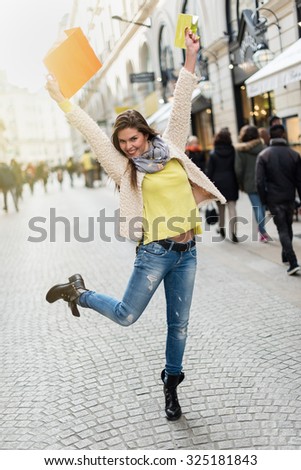Smiling woman in her 30s with black boots and white coat jumping in the air, she is raising her shopping bags  to show her happiness. She is in a car-free street in the city center.