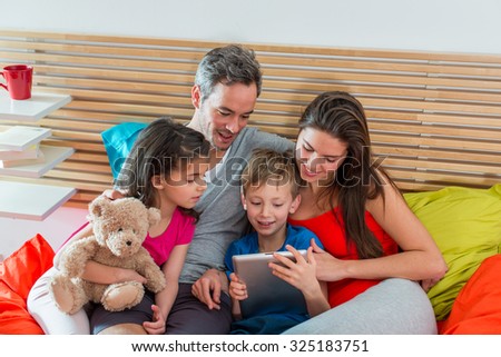 Parents and their two children are sitting in bed in the morning. They are watching tablet. The little girl is holding her cuddly toy. There is a wooden headboard behind them
