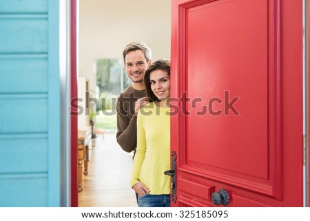 A grey hair man with beard and a woman are opening the red door of their wooden house to welcome some guests. The thirty years old couple is smiling, wearing sweater and jeans.