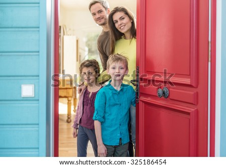A nice four people family is opening their stylish red door to welcome the guest. The parents and their two children are smiling and wearing casual clothes. The house have blue wall and wooden floors