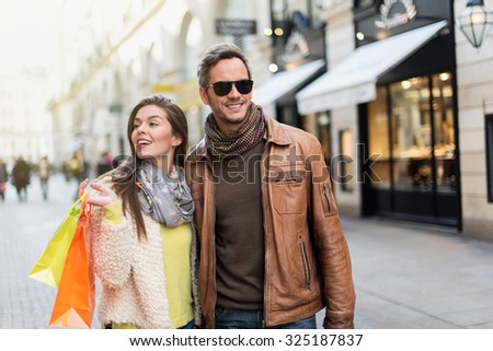 Stylish couple walking in a cobbled car-free street. The grey hair man with beard is wearing sunglasses and a brown leather coat and the woman a yellow top and two shopping bags, they also have scarfs