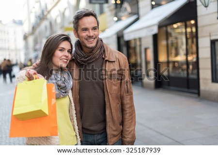 Stylish couple standing in a cobbled car-free street. The grey hair man with beard is wearing a brown leather coat and the woman a yellow top and two shopping bags, they also have scarfs