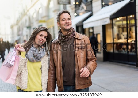 A trendy couple is walking in the city center. They are in a cobbled car-free street. The woman is wearing a yellow shirt and pink shopping bags and the grey hair man with beard has a leather coat
