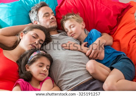 A family is taking a nap all together in a colorful bed with large cushions. The mother and the two children have lay their heads on the gray hair father. They look peaceful with their eyes closed