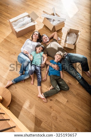 Top view of a happy family in casual clothes laying on the wooden floor of their new flat with cardboard boxes around them They are looking at camera The smiling parents are holding their two children