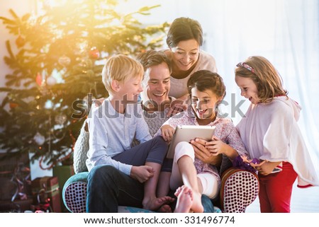 Christmas morning, cheerful family sitting in the living room having fun with the digital tablet that Santa Claus brought her, behind the decorated christmas tree, the sunshine give a cozy atmosphere