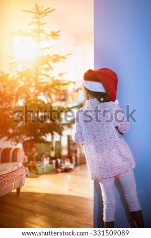 Christmas morning, a little girl in pajamas, wearing a Santa Claus hat opens the living room door and discovers the Christmas tree and many gift.The sun coming through the window gives cozy atmosphere