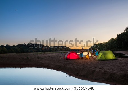 Group of teenagers having fun at sunset on the beach. They are camping with two red and green tents They are playing guitar and dancing around a camp fire. They are wearing summer hats and sunglasses