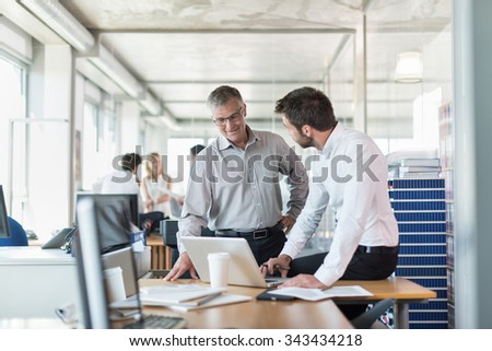 Two senior partners standing in front of a desk in a luminous open space with glass walls. They are analyzing balance sheets on a laptop. They are wearing a shirt and suit pants.