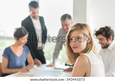 Portrait of a smiling blond assistant, she is turned back on her chair at an office business meeting. The team is sitting at a table in a luminous white open space, brainstorming some new ideas.