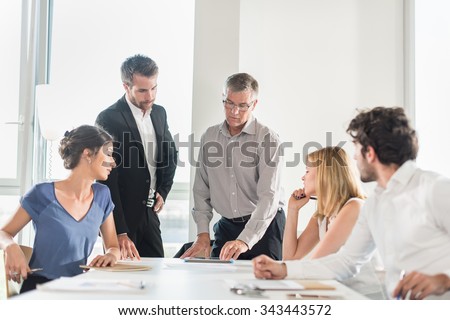 Office business meeting. The team is sitting at a table in a luminous white open space The men are wearing suits and shirts. The senior grey haired boss is up and comparing a new design on his tablet