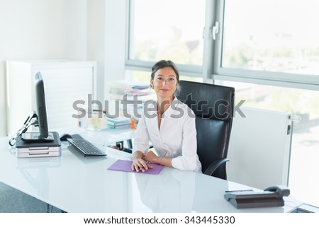 Smiling manager looking at camera in her white office Woman sitting at a tidy glass desk in front of a black computer She is wearing a white shirt her hair are tied Her hands are crossed on a folder