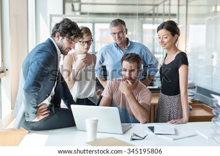 Office meeting, everybody is looking at the computer engineer website presentation. He is sitting at a desk in a luminous open space, the team is standing around him with the grey hair senior partner
