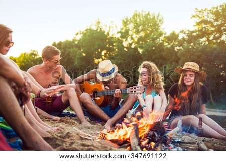 Teenagers having fun at the beach. They are sitting in the sand in circle around a camp fire, wearing short pants, sunglasses and straw hats. They are singing and a boy is playing guitar