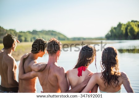 Back view of a group of teenagers looking at the horizon. They are standing arm in arm, like best friends, in front of an amazing river with her feet in the water. They are wearing swimsuits