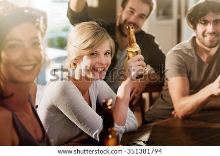 Backlit shot with flare. Friends having a drinks on a sunny evening in a bar, They are sitting with beers. They are wearing casual clothes. Focus on a gorgeous blonde girl touching her bottle.