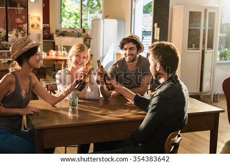 Four friends in their 30\'s having a drink after work. Two men and women are sitting at a wooden table joining their bottles of beer in a cozy house. They are smiling, wearing casual clothes and hats.