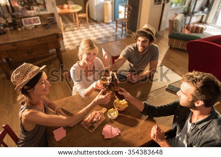 Top view of friends having a drink and playing poker on a sunny evening. They are sitting at a wooden table in a cozy house with beers and tortilla chips. They are wearing casual clothes and hats.
