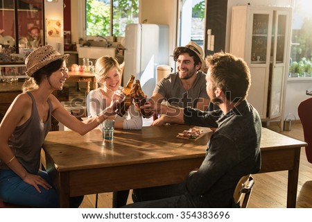 Four friends in their 30's having a drink after work. Two men and women are sitting at a wooden table joining their bottles of beer in a cozy house. They are smiling, wearing casual clothes and hats.