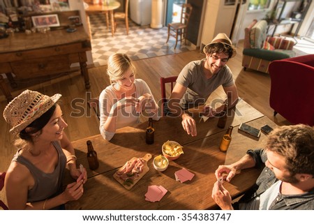 Top view of friends having a drink and playing cards on a sunny evening. They are sitting at a wooden table in a cozy house with beers and tortilla chips. They are wearing casual clothes and hats.