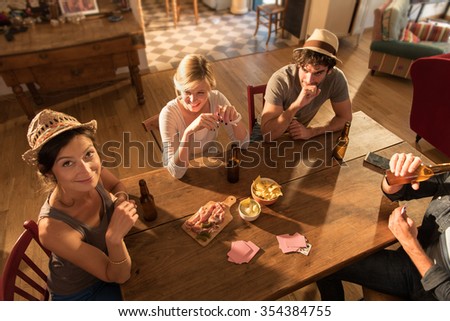 Top view of friends having a drink and playing cards on a sunny evening. They are sitting at a wooden table in a cozy house with beers and tortilla chips. They are wearing casual clothes and hats.