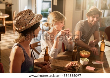 Friends having a drink and playing cards on a sunny evening. They are sitting at a wooden table in a cozy house with beers and tortilla chips. They are smiling, wearing casual clothes and hats.