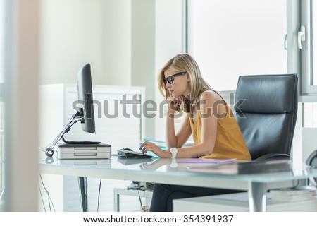Portrait of a blonde female business partner in her 30\'s sitting at her tidy glass desk and typing on the keyboard of her computer in a luminous white office. She is wearing a yellow top and glasses.