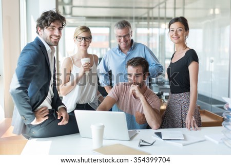 Real people business portrait, everybody is looking at camera. The computer engineer is sitting at a desk in a luminous open space and the team is standing around him with the grey hair senior partner