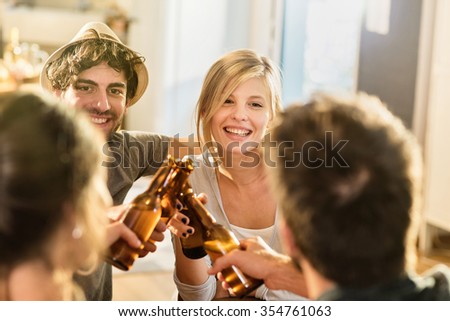 Friends having a drinks on a sunny evening in a cozy house. They are sitting at a wooden table. They are joining their beers. They are wearing casual clothes. Focus on a nice couple in his 30\'s.