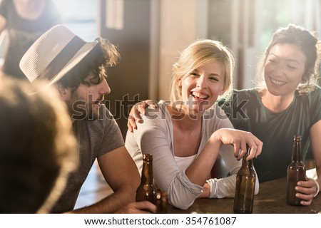 Friends having a drinks on a sunny evening in a cozy house. They are sitting at a wooden table with beers. They are wearing casual clothes. Focus on a gorgeous blonde girl touching her bottle.