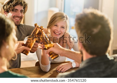 Friends having a drinks on a sunny evening in a cozy house. They are sitting at a wooden table. They are joining their beers. They are wearing casual clothes. Focus on a gorgeous blonde girl.