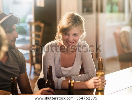 Friends having a drinks on a sunny evening in a cozy house. They are sitting at a wooden table with beers. They are wearing casual clothes. Focus on a smiling blonde girl holding her bottle.