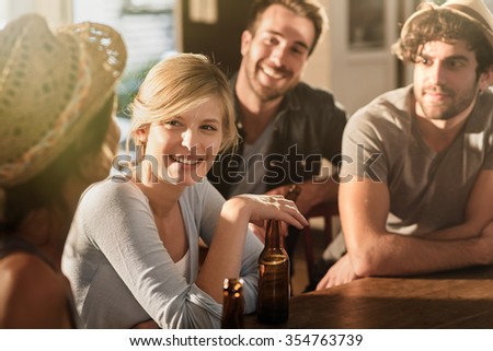 Backlit shot with flare. Friends having a drinks on a sunny evening in a bar, They are sitting at a wooden table with beers.Focus on a gorgeous blonde girl touching her bottle.