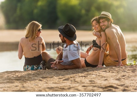 Back view of two young couple in their 30\'s at the beach. They are sitting in the sand arm in arm, enjoying the last lights of the day and looking at the ocean. They are wearing swimsuits and hats.