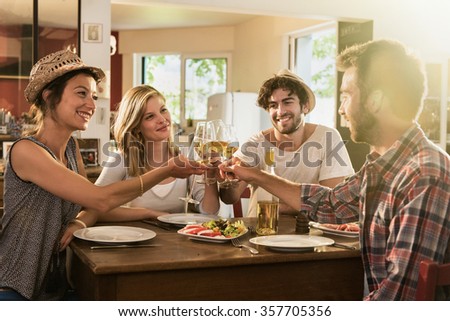 Friends in their 30s having a nice aperitif on a rustic wooden table in a lovely house. They are holding their high glasses of white wine. There are tomatoes mozzarella for starters. Shot with flare