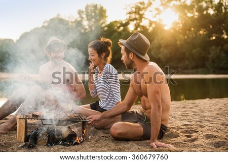 group of friends having fun at the beach. They are sitting around a campfire with a lot of smoke, doing a barbecue with skewers  A shirtless man with beard and hat is playing a ukulele.