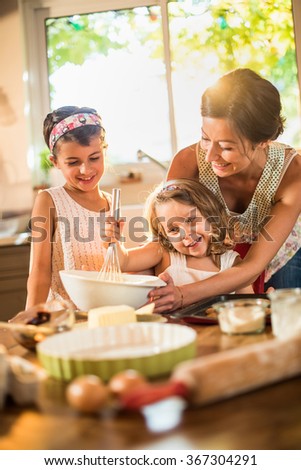 A mother is cooking with her two daughter of four and seven years old. They are smiling, wearing casual clothes. The woman is helping her youngest mixing the ingredients in a large bowl.