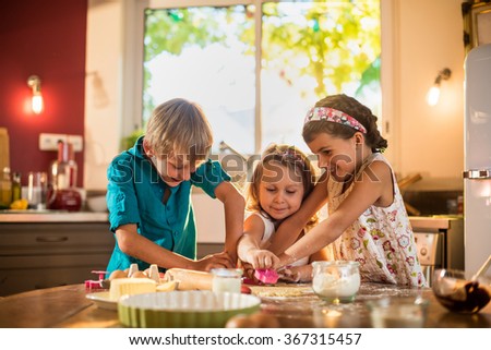 Three brother and sisters are cooking together in a luminous kitchen. They are standing close to each other at a wooden table, working on a spread pastry in order to make small cakes. Shot with flare