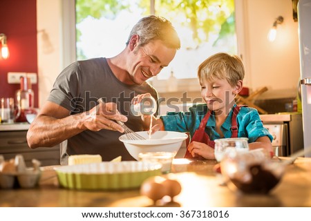 A eight years old blond boy is cooking  with his father in a luminous kitchen. They are sitting at a wooden table the dad is mixing the preparation while his son is adding ingredients.Shot with flare