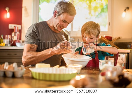 A eight years old blond boy is cooking  with his father in a luminous kitchen. They are sitting at a wooden table the dad is mixing the preparation while his son is adding ingredients.Shot with flare