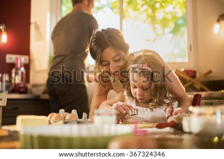 A mother and her four years old blonde daughter are cooking in a luminous kitchen. They are making forms on a pasty on a wooden table full of ingredients. The little girl is focused on her actions.
