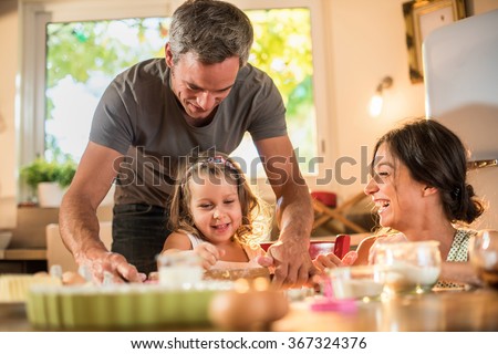 A four years old blonde girl is cooking small cakes with her mother and grey haired father in a luminous kitchen. They are working on a pastry with a rolling pin on a wooden table full of ingredients