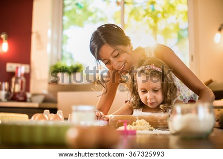 A mother and her four years old blonde daughter are cooking in a luminous kitchen. They are working on a pastry with a rolling pin on a wooden table full of ingredients and a green baking pan.