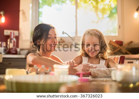 A mother and her four years old blonde daughter are cooking in a luminous kitchen. They are having fun, working on a pasty on a wooden table full of ingredients and a baking pan. Shot with flare