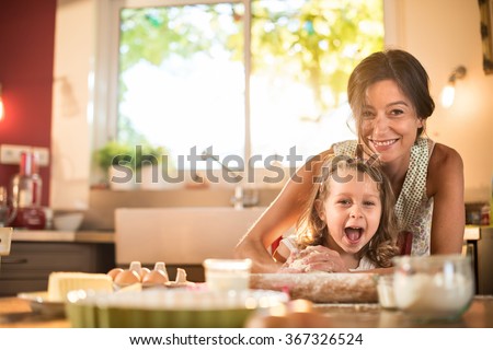 A mother and her four years old blonde daughter are cooking in a luminous kitchen. They are having fun, working on a pastry with a rolling pin on a wooden table full of ingredients and a baking pan.